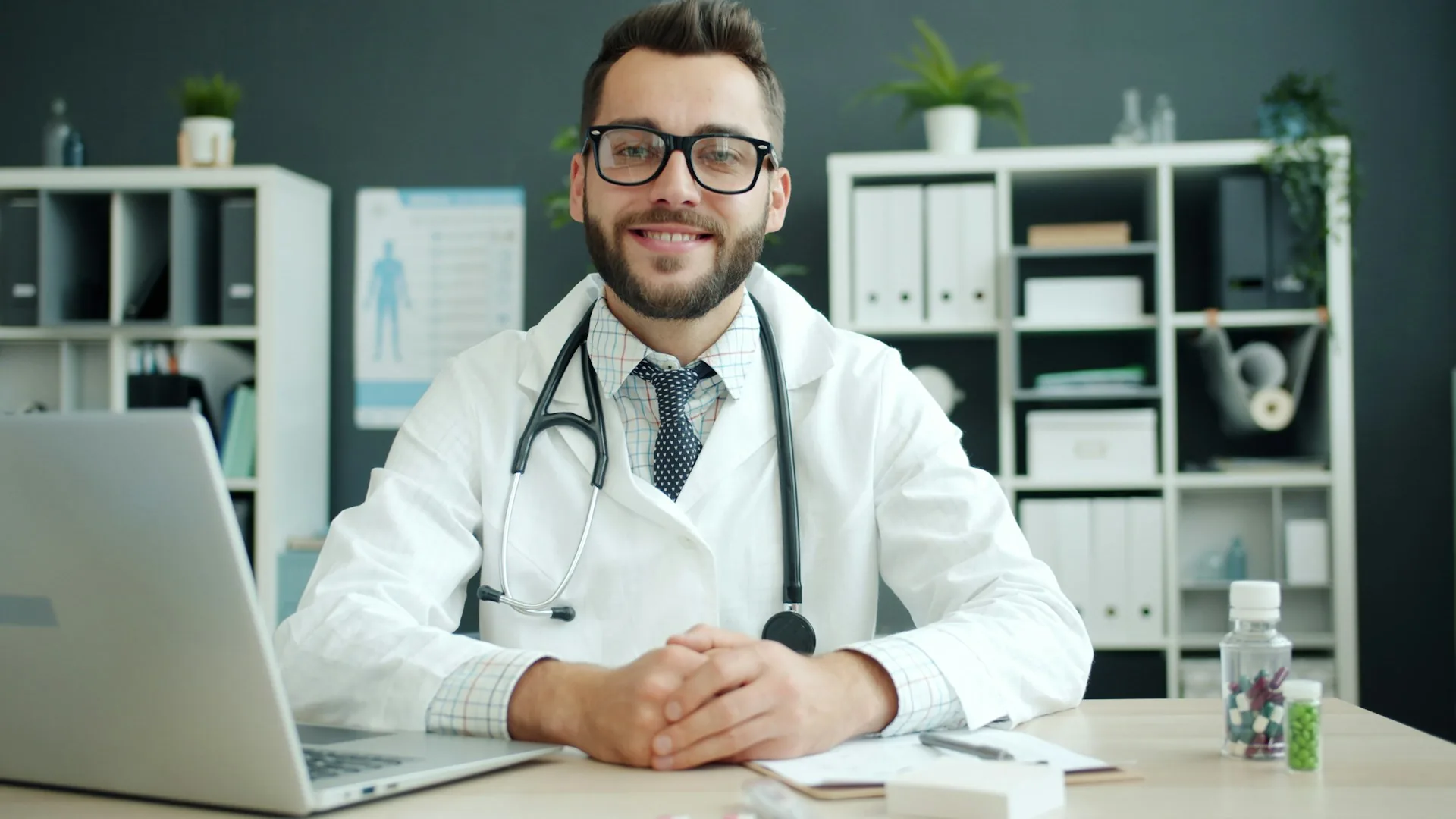 Médico con bata blanca y fonendoscopio sentado en su consulta mirando a cámara